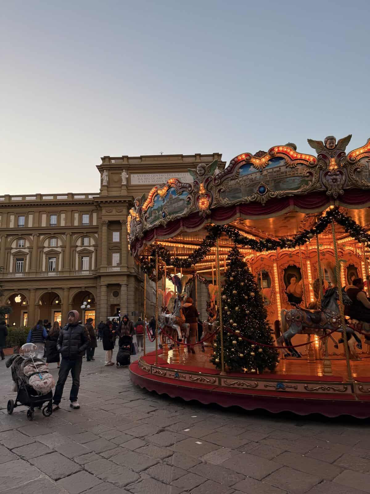 An ornate, illuminated carousel decorated with garlands and a Christmas tree in a city square, with a man standing nearby with a stroller during 3 days in Florence.