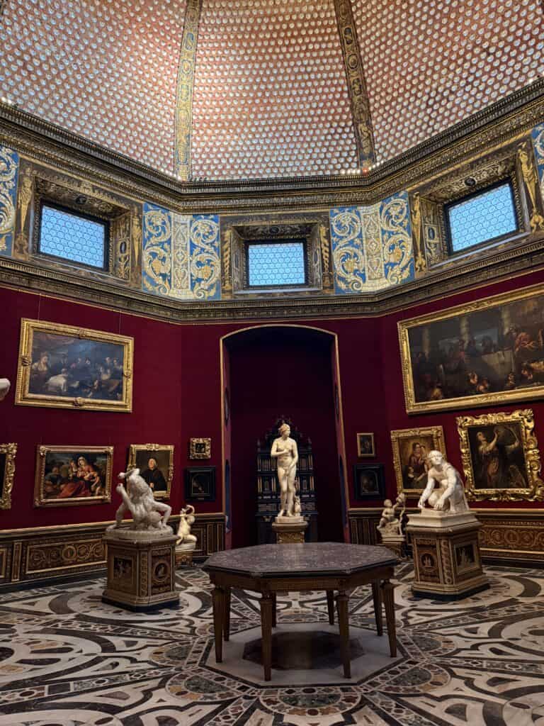 The Tribuna room at the Uffizi Gallery, a highlight for any 3 days in Florence, features Renaissance paintings on red walls, classical statues, and a central octagonal table under a domed ceiling.